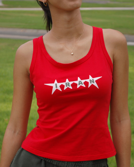 A woman wearing a bright red tank top with a white graphic that spells 'IRAN' across four stars, photographed outdoors against a blurred green grass background.