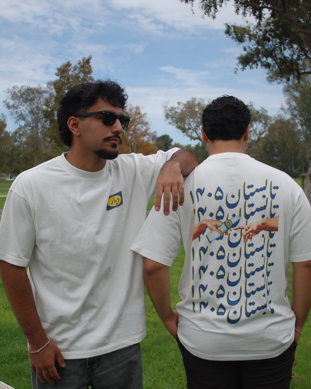 Two men standing outdoors in a park wearing white graphic t-shirts. The man on the left is shown from the front with a small yellow and blue chest logo, while the man on the right is shown from the back with a large blue and yellow graphic print featuring Farsi calligraphy and hands holding a glass bottle of doogh.